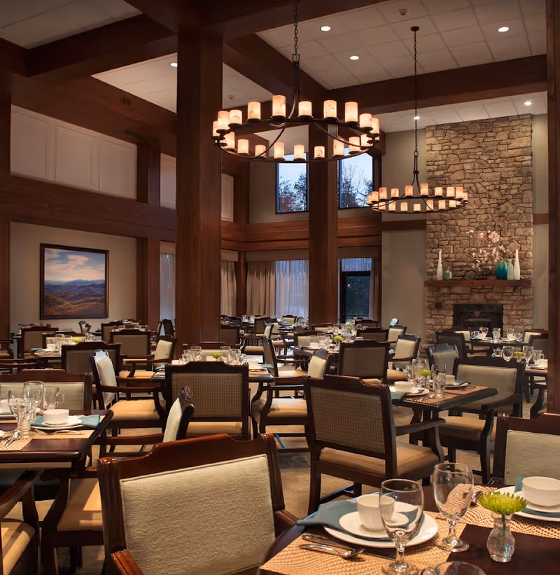 A spacious dining room with multiple tables set with plates, glasses, and silverware. The room features large wooden beams, two circular chandeliers with candle-like lights, a stone fireplace with decorative vases, and large windows with curtains. A framed landscape painting is visible on one wall.
