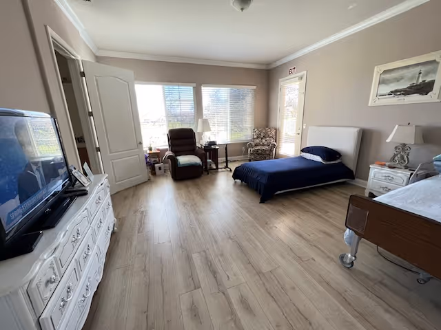 Bright senior living bedroom with a single bed, dresser, recliner, and large windows.