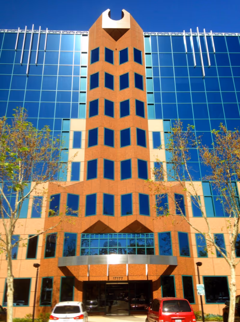 Glass-fronted modern building with brick central tower, trees, and cars at the entrance.