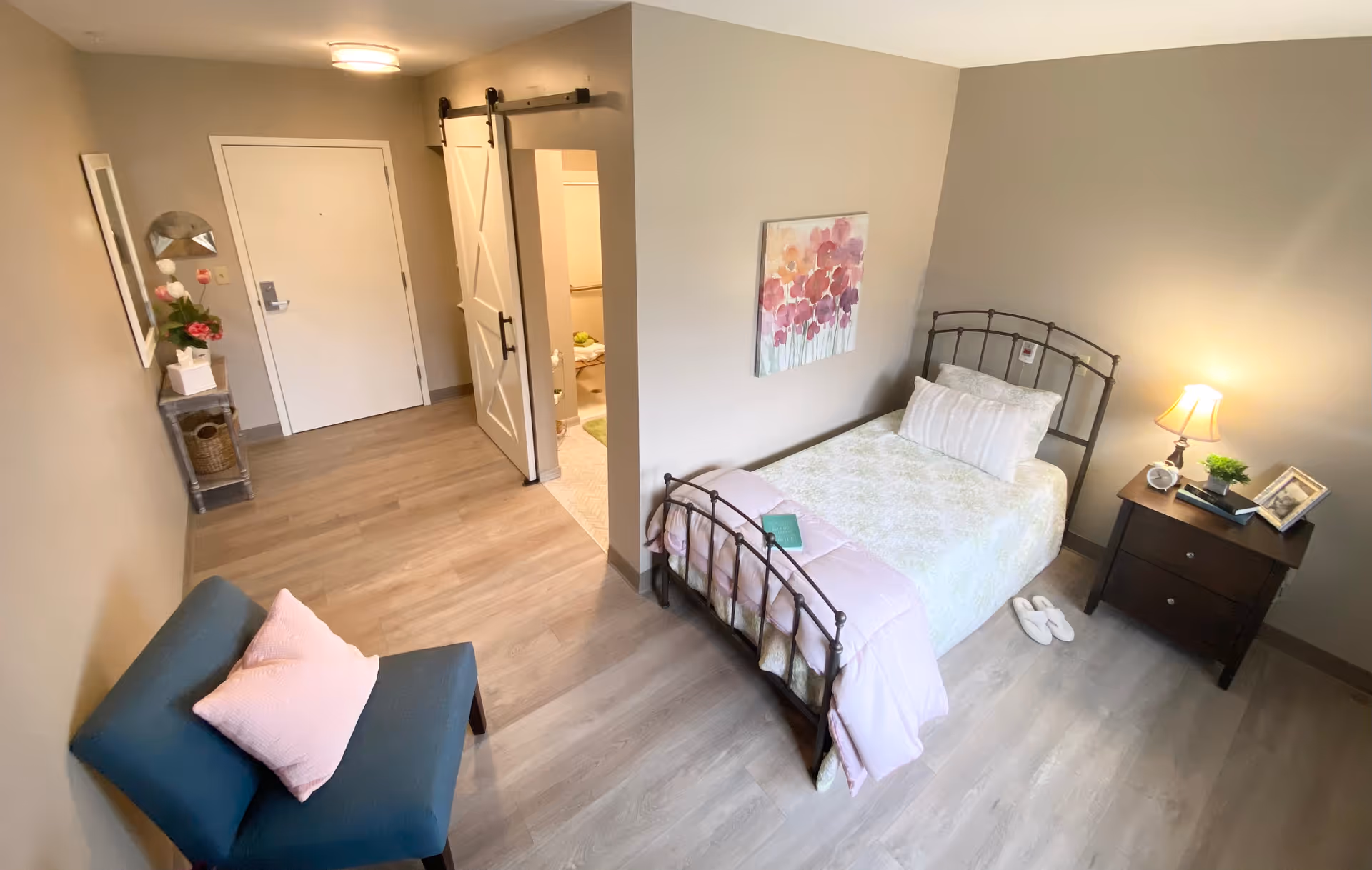 Cozy resident bedroom with a metal bed, nightstand, chair, and doorway to hallway.