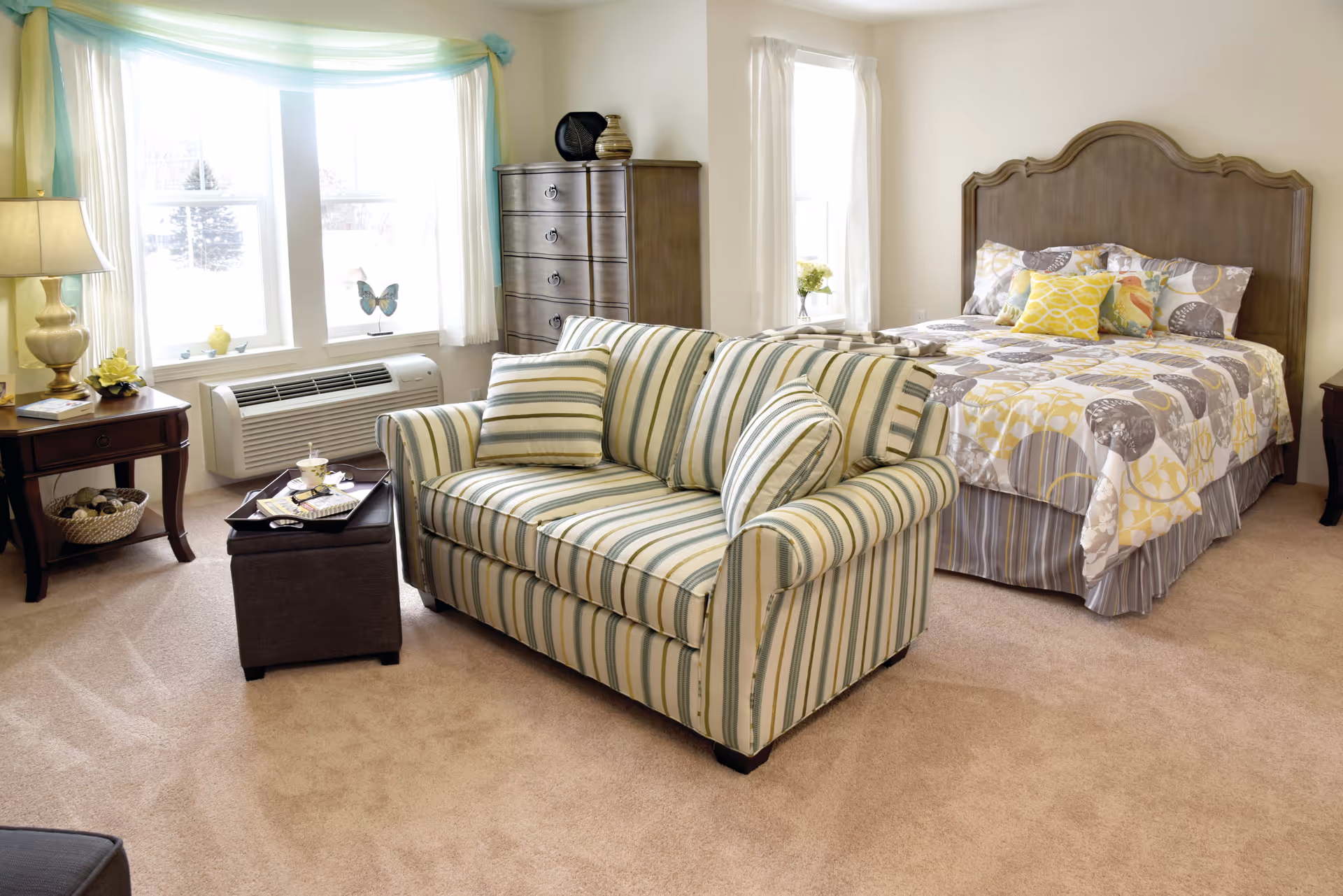 A bright and cozy bedroom featuring a wooden bed with patterned yellow and gray bedding, a striped loveseat with matching pillows, a wooden dresser, and a side table with a lamp and decorative items. Large windows with white curtains and light blue and yellow valances allow natural light to fill the room.