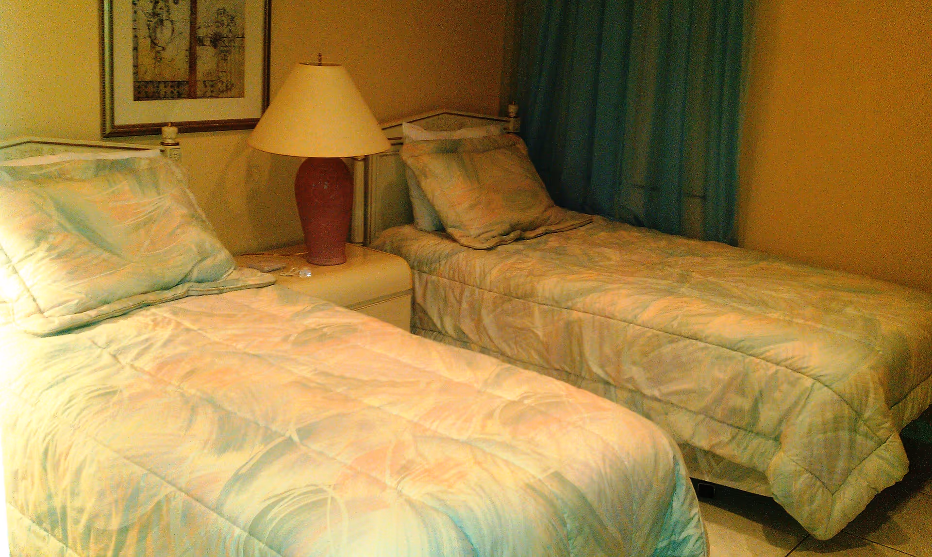 A bedroom with two single beds placed side by side, each with pastel-colored bedding and pillows. Between the beds is a nightstand with a red lamp. A framed picture hangs on the wall above the nightstand, and a blue curtain covers the window on the right side of the room.