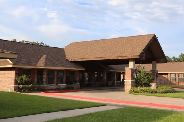 Front entrance of a single-story brick nursing facility with a covered porte-cochere, driveway, and landscaped lawn.