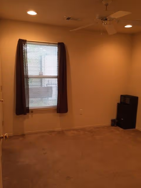 Empty beige room with window, dark curtains, ceiling fan, and a speaker on carpet.