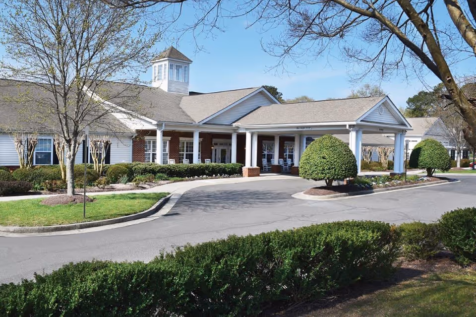 Exterior view of a senior living facility building with a covered entrance, white columns, and a small cupola on the roof. The building is surrounded by neatly trimmed bushes, trees, and a curved driveway under a clear blue sky.
