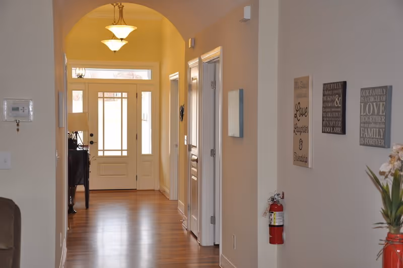 A well-lit hallway with wooden flooring leading to a front door with glass panels. The hallway has an arched ceiling with two hanging light fixtures. On the right wall, there are three decorative wall plaques with inspirational quotes and a fire extinguisher mounted below them. A small table with a lamp is visible on the left side near the door.