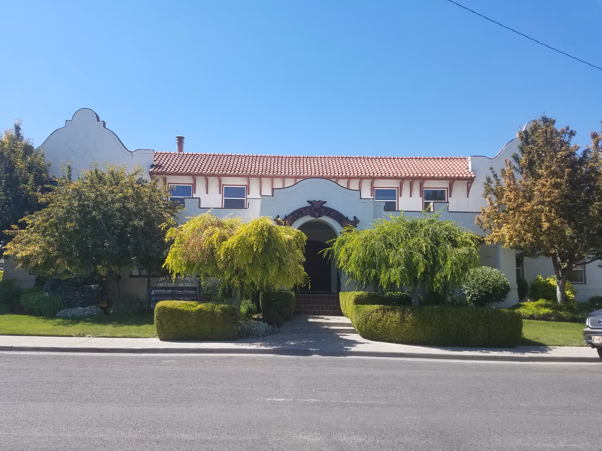 Beige stucco senior living building with a red tile roof and trimmed trees by the entrance.