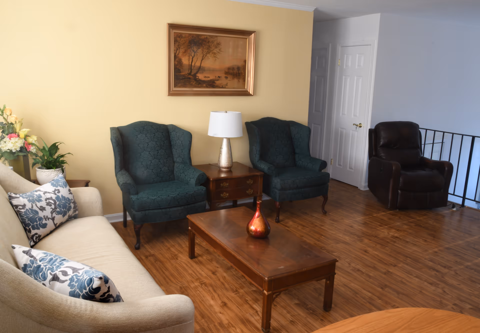 A cozy living room with a beige sofa adorned with patterned blue and white pillows, two dark green upholstered armchairs, a wooden coffee table with a decorative red vase, a wooden side table with a lamp, a framed landscape painting on a yellow wall, and a dark brown leather recliner near a white door and black railing.