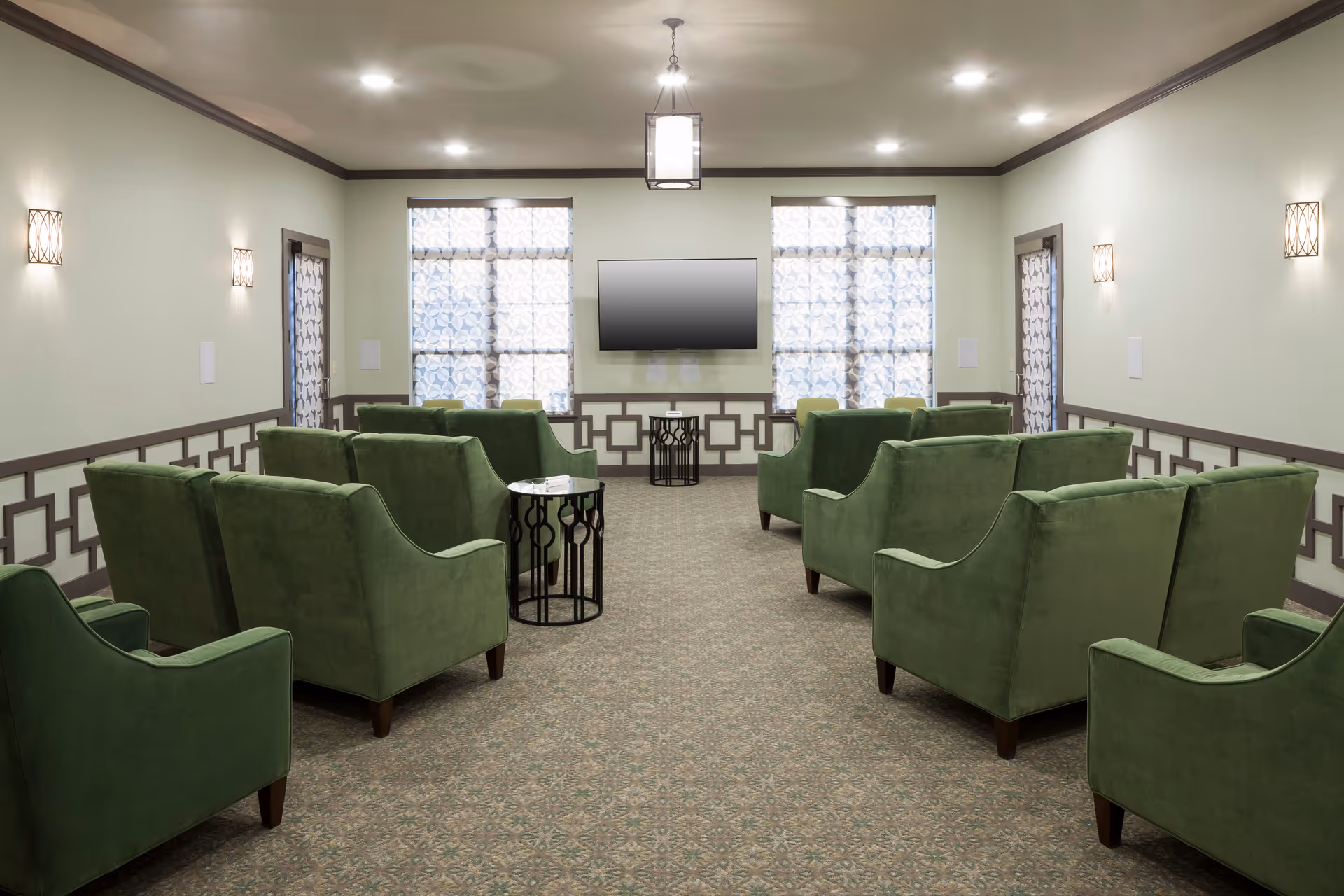 Spacious lounge area with green armchairs arranged around small tables facing a wall-mounted TV.