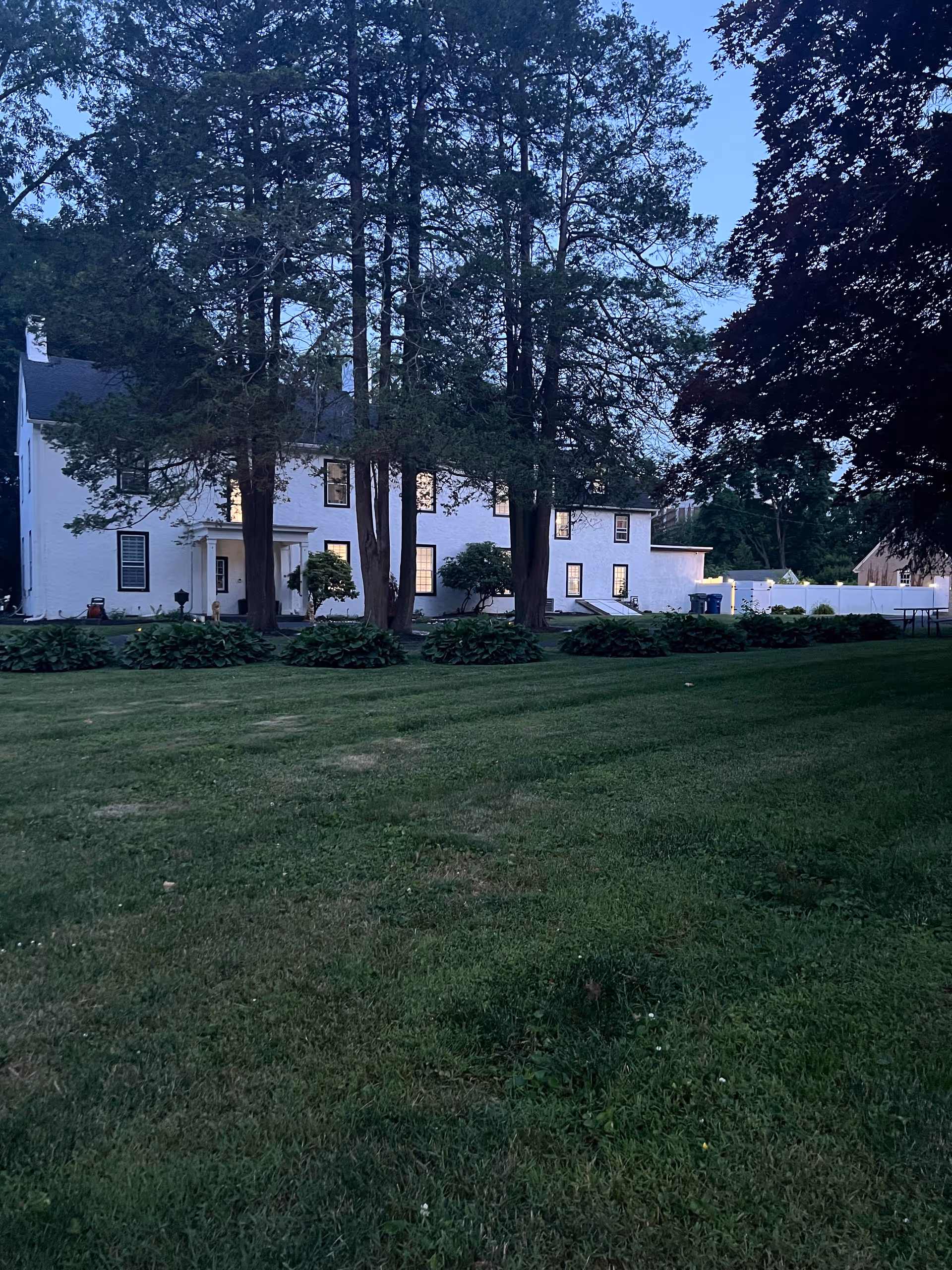 A white two-story senior care home with tall trees and a manicured front lawn.