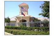 Front view of a beige senior living facility with a tower, flags, and a fence.