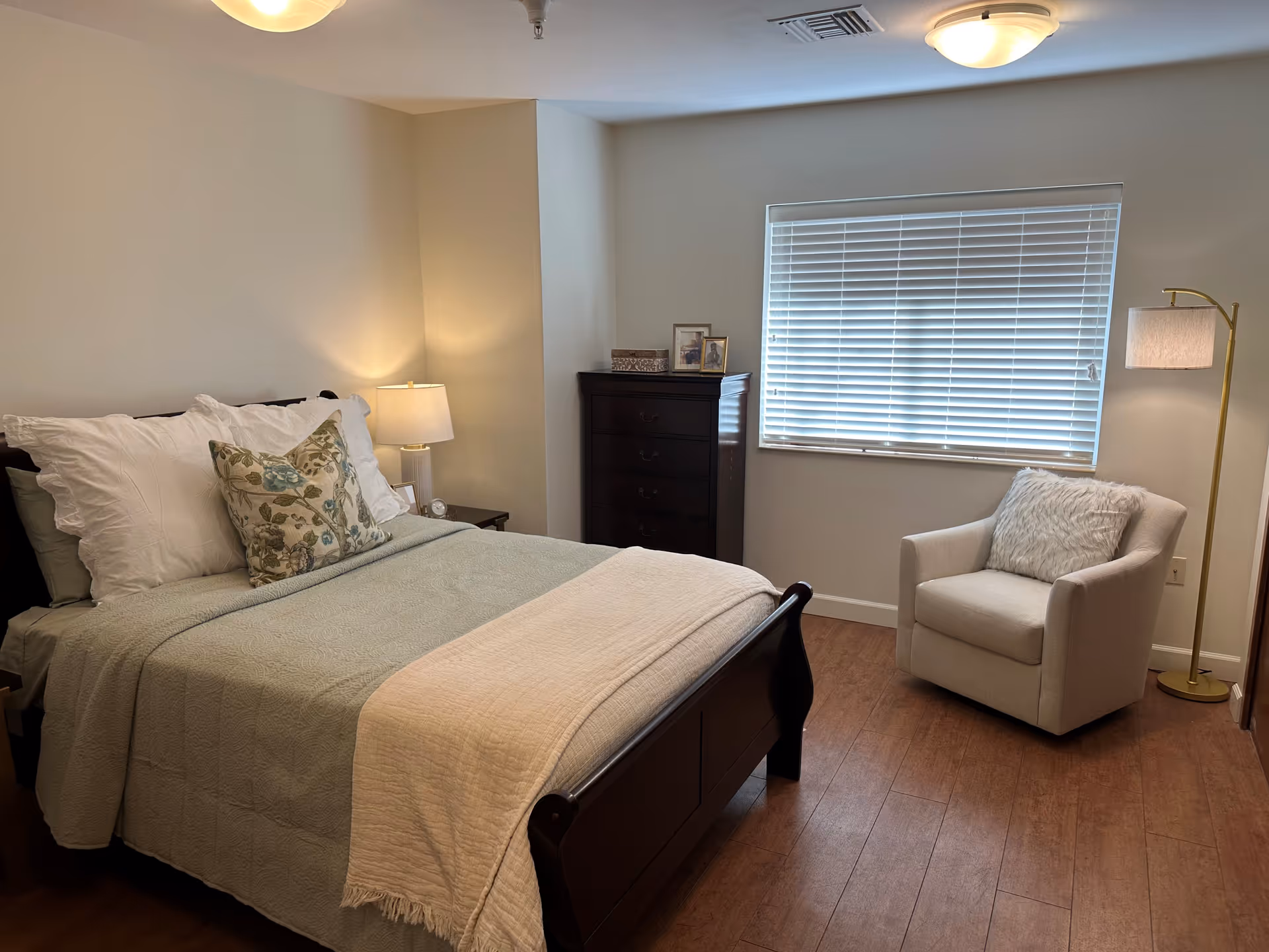 A cozy bedroom with a wooden bed frame, neatly made with white and floral pillows and a light green quilt. There is a wooden nightstand with a lamp on the left side of the bed. A tall dark wooden dresser is placed against the wall near a window with white blinds. To the right of the window, there is a beige armchair with a fluffy pillow and a tall floor lamp with a white shade. The room has wooden flooring and neutral-colored walls.