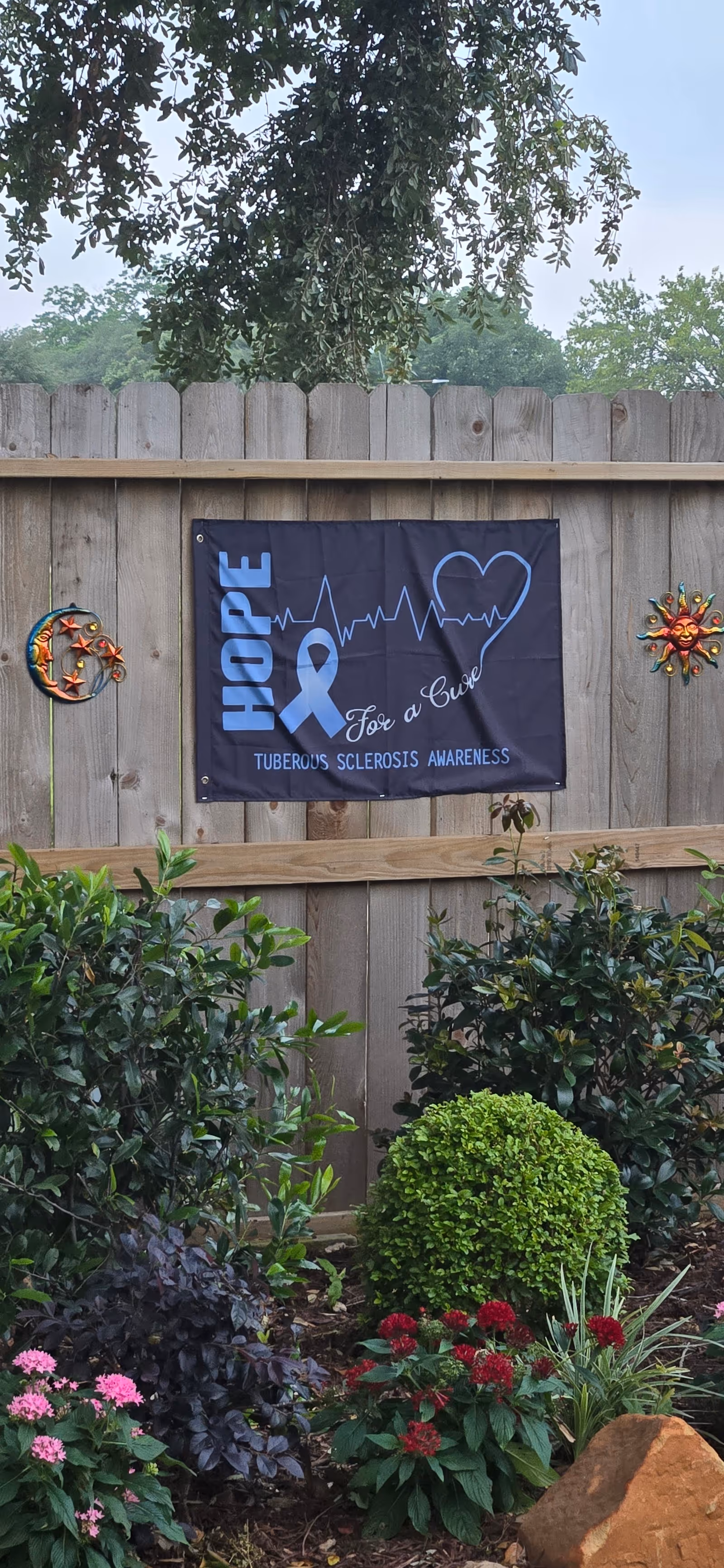 Wooden fence displays a Hope For a Cure banner with a garden in front.