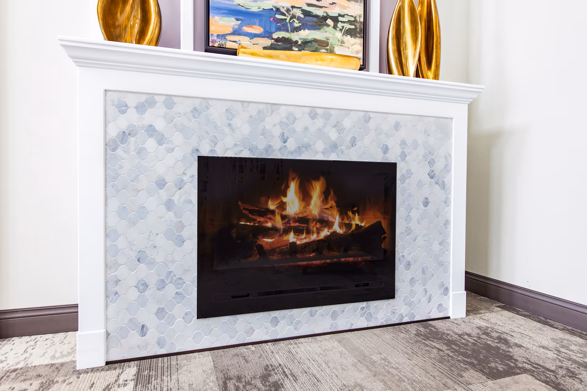 Modern fireplace with a burning fire set in a white mantel with a light marble mosaic surround and decorative items on the mantel.