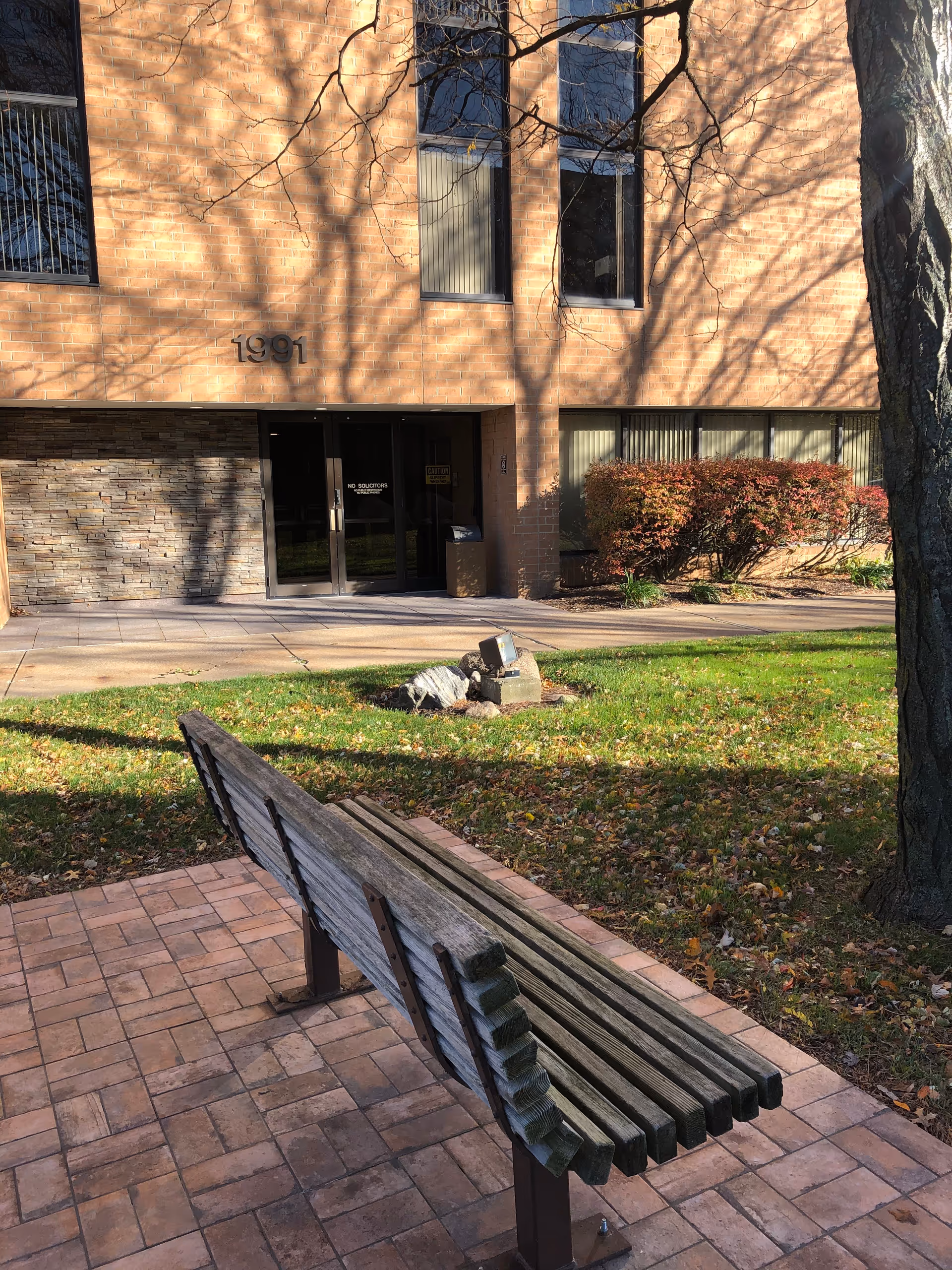 Brick senior living building entrance with benches, trees, and a shaded walkway.