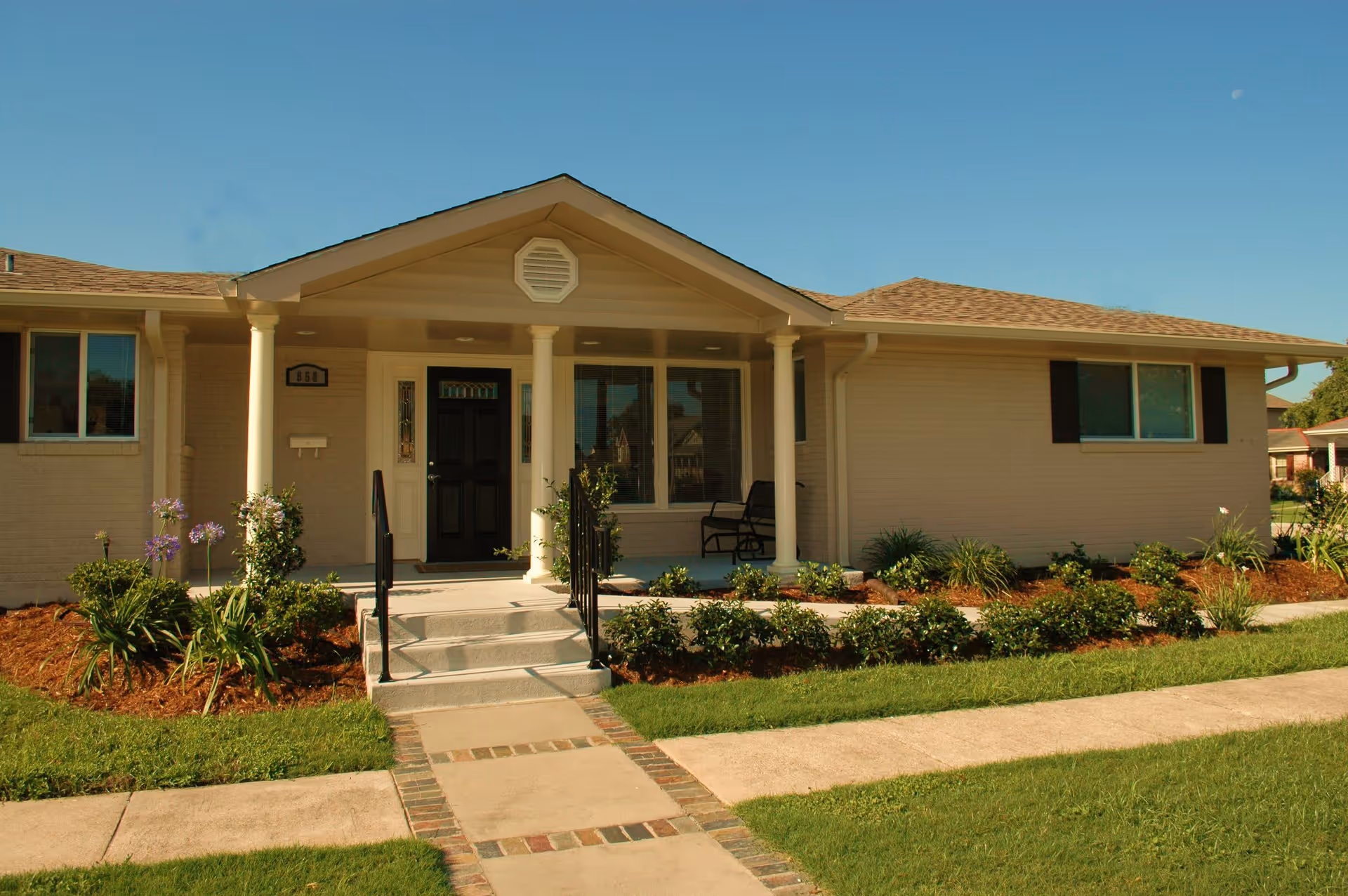 Single-story light-colored ranch-style house with a front porch, steps, columns, and a landscaped yard under a clear blue sky.