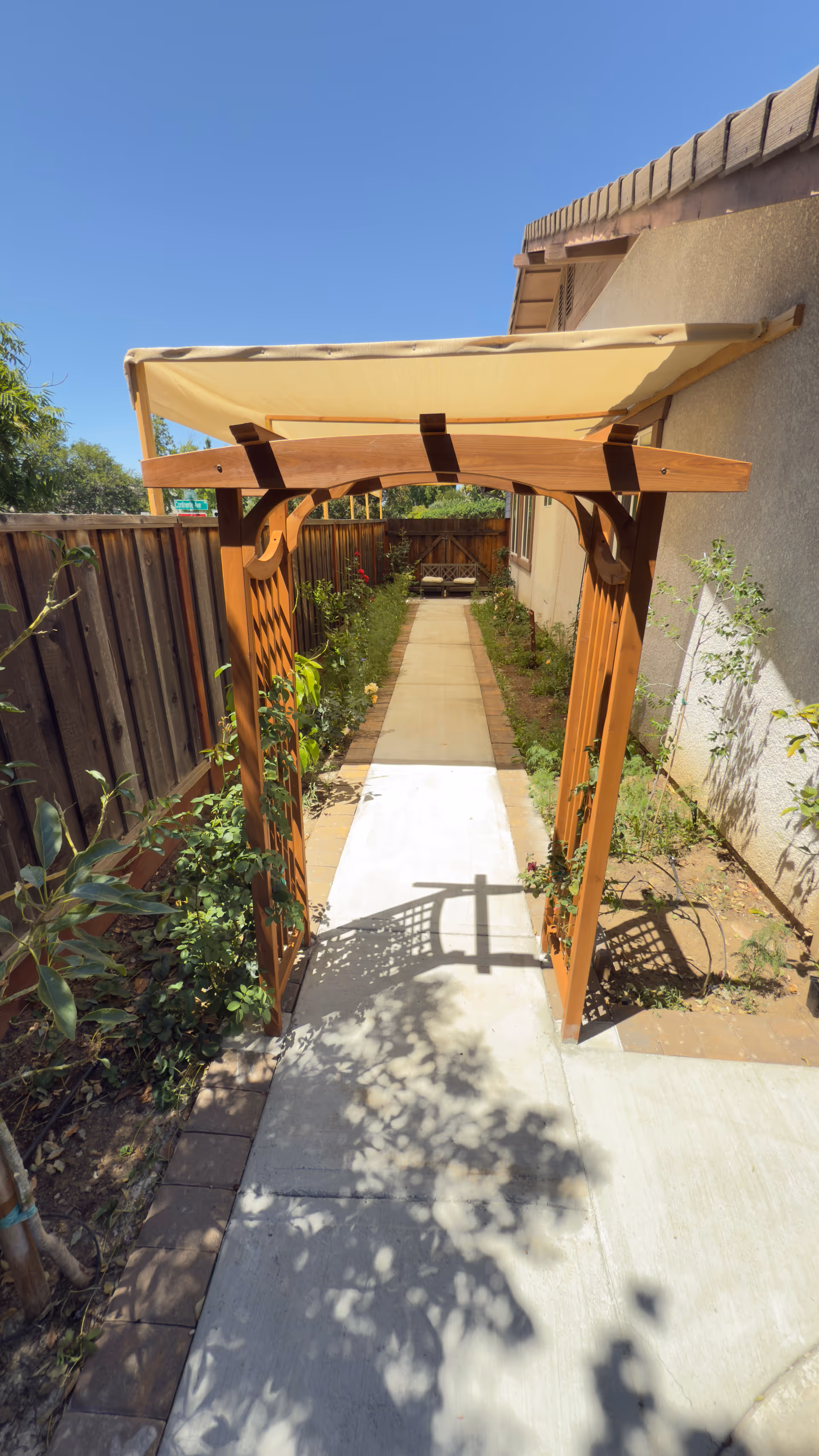 Wooden pergola archway over a concrete path beside a house in a fenced garden.
