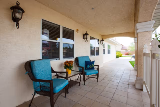 Covered front porch with two wicker chairs with blue cushions, a small table with flowers, wall lanterns, and a tiled walkway.