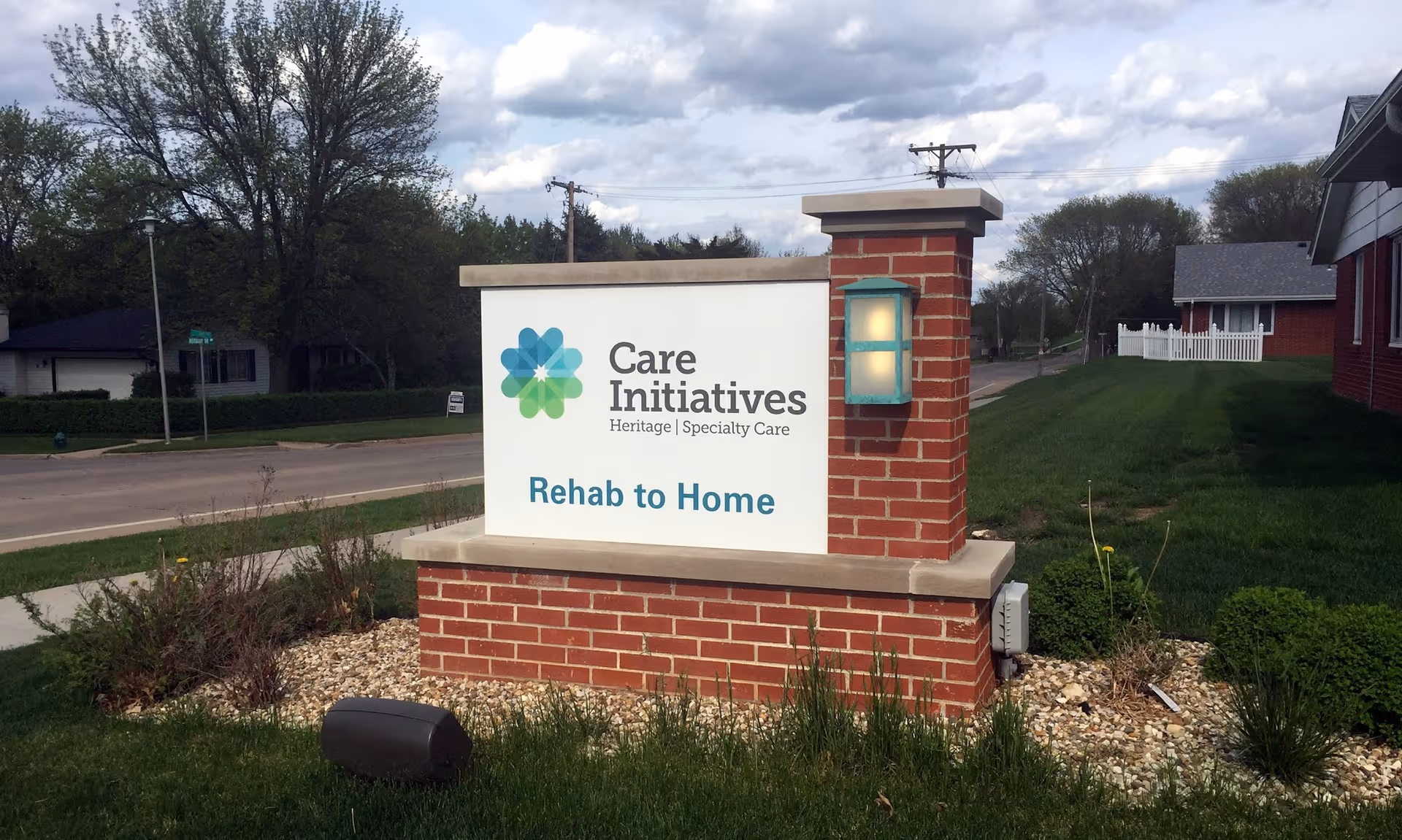 Brick monument sign reading Care Initiatives Heritage | Specialty Care Rehab to Home on a grassy lawn by a street.
