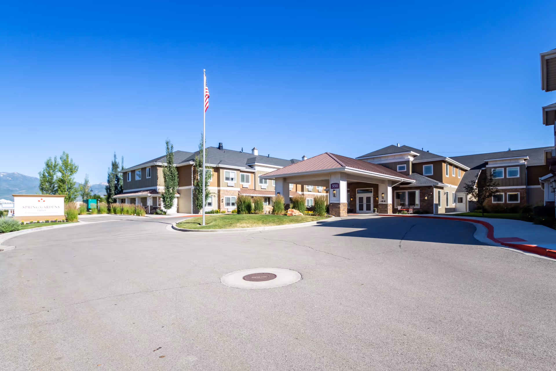 Exterior view of Spring Gardens Senior Living Heber facility on a clear sunny day, showing the main entrance with a covered drop-off area, an American flag on a flagpole, and surrounding buildings with landscaping and a paved driveway.