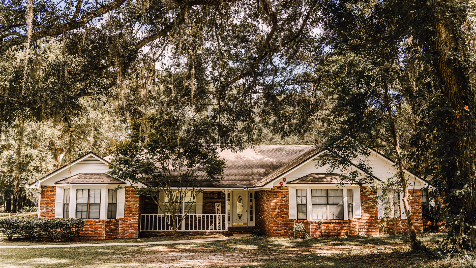Single-story brick house with a front porch, surrounded by trees and a lawn.