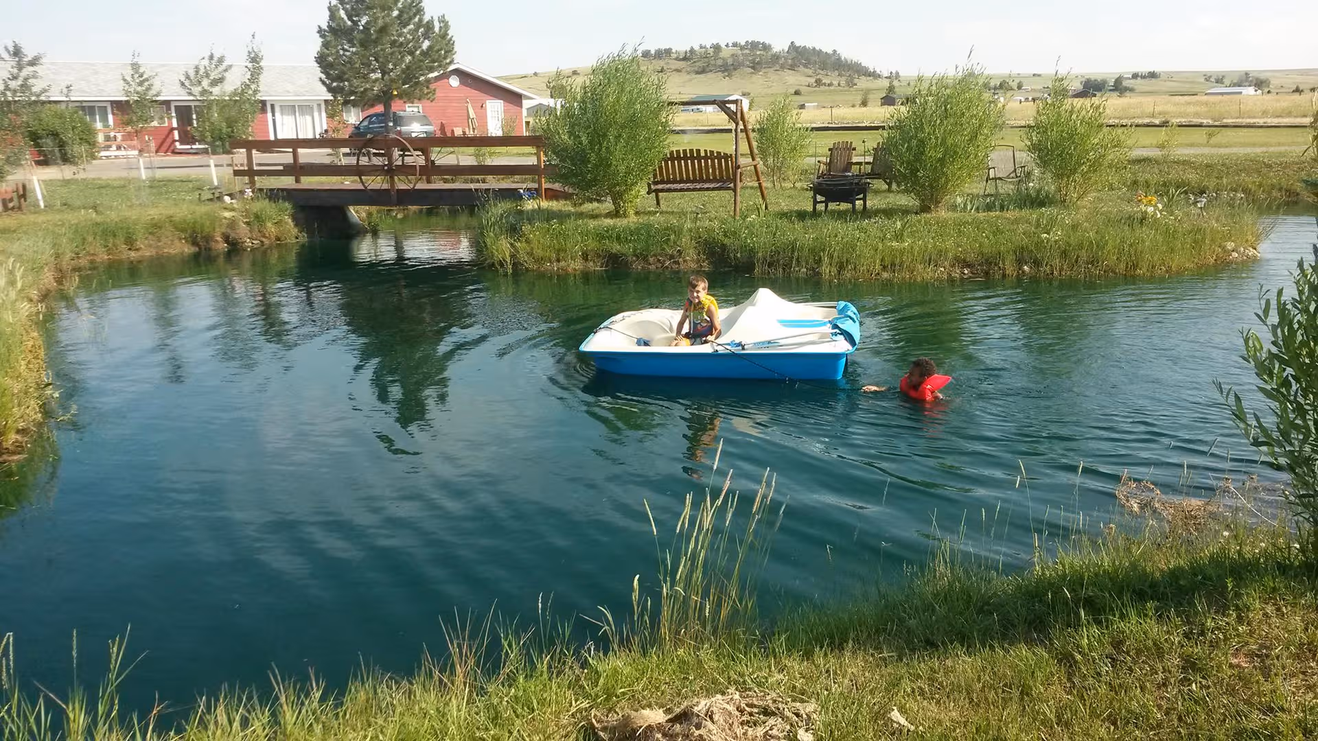 A pond with a blue boat carrying a child, near a wooden bench and red building.