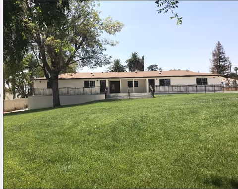 Single-story care facility building with a wide green lawn, trees, and a ramped entrance.