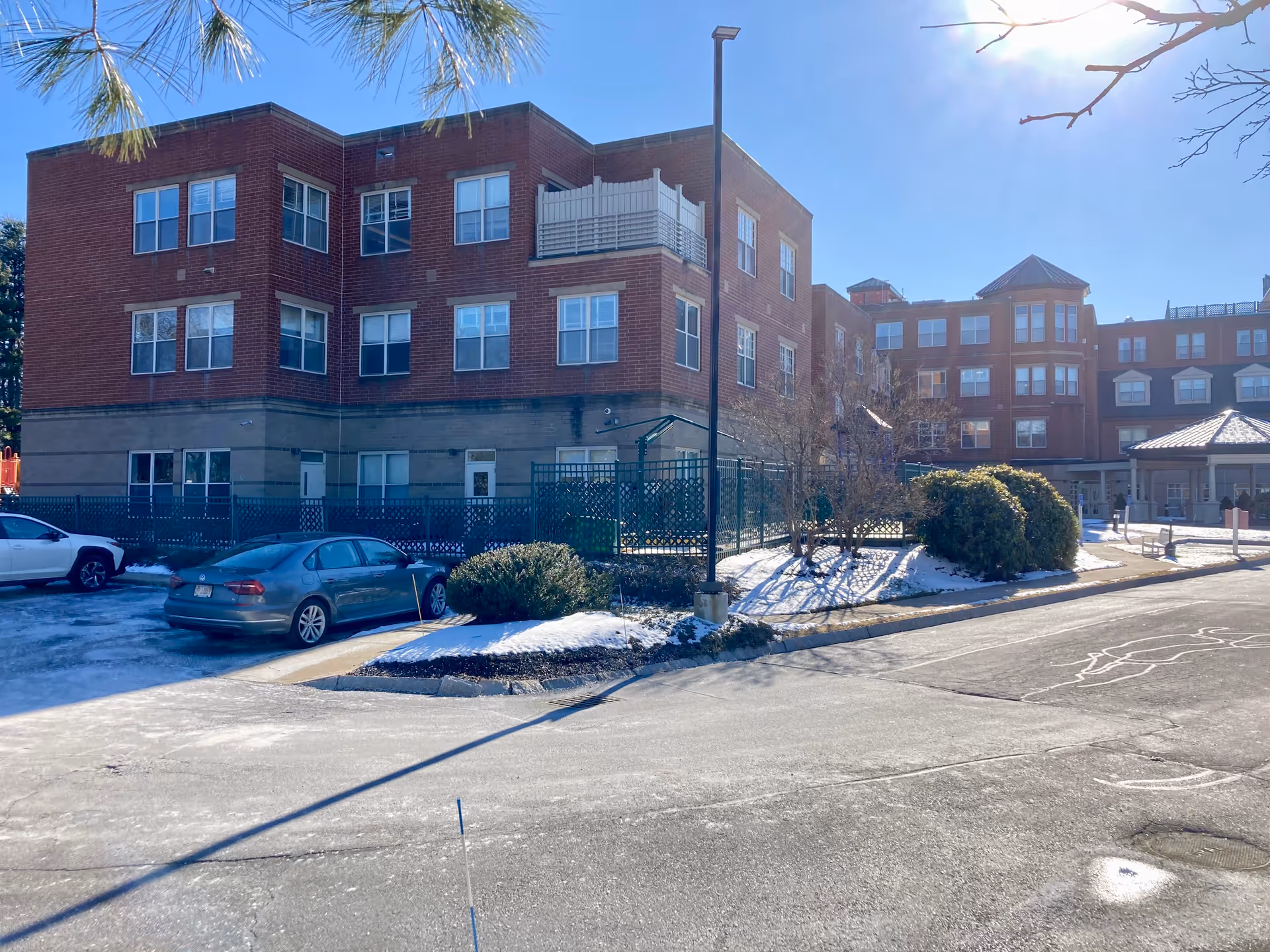 Exterior view of a multi-story brick building with several windows, a small parking area with cars, some bushes, and patches of snow on the ground under a clear blue sky.