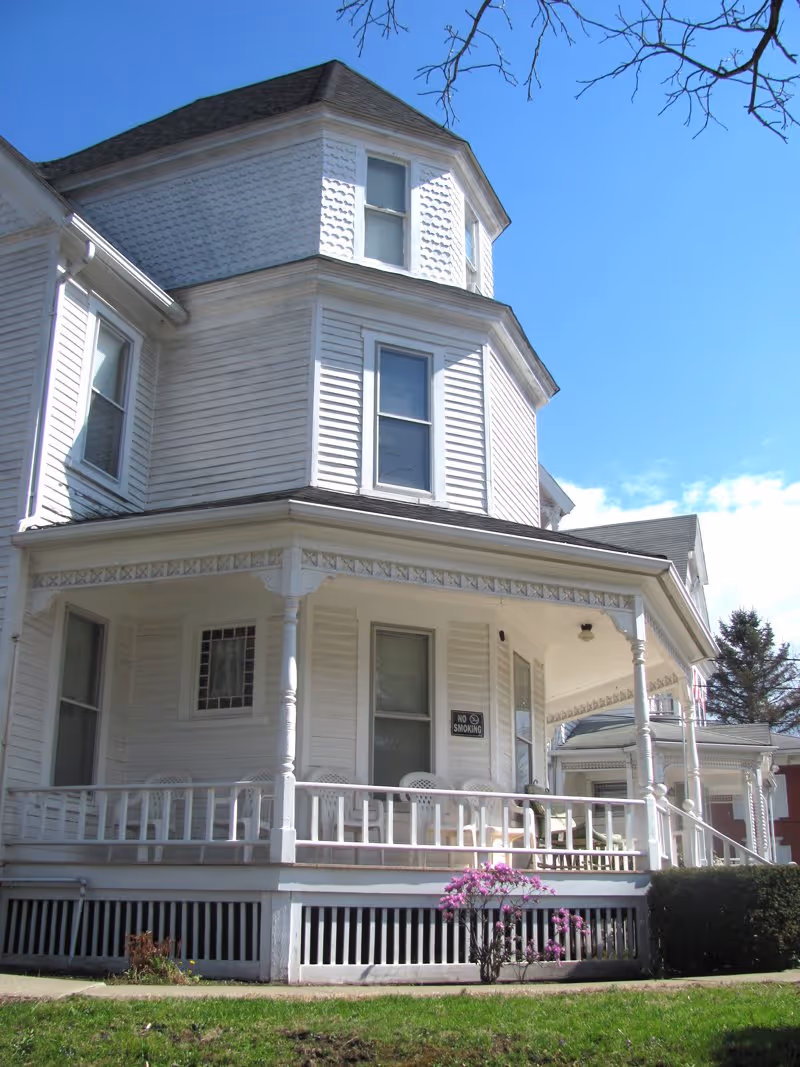 White Victorian-style house with a wraparound porch, chairs, and bright blue sky.
