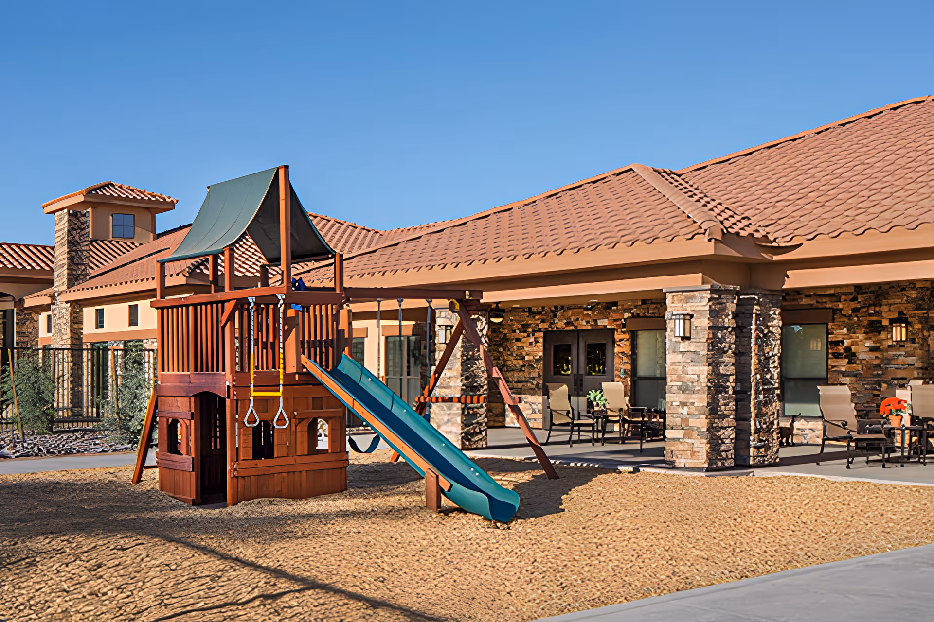 A wooden playground with a green slide and swings sits in a sandy courtyard in front of a single-story building with stone columns and a red tiled roof.