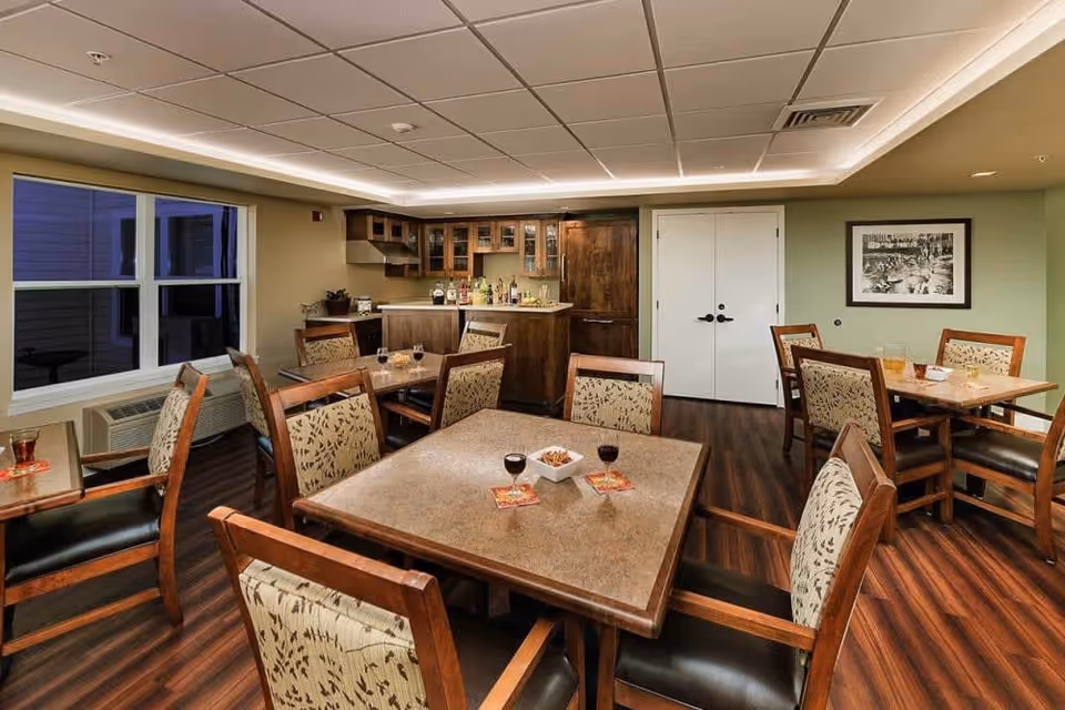 Interior view of a dining room in a senior living facility with several wooden tables and cushioned chairs arranged neatly. The room features a small bar area with cabinets and a countertop stocked with bottles. The floor is wooden, and the ceiling has recessed lighting. A window on the left side shows an exterior view, and a framed black and white photograph hangs on the wall to the right.