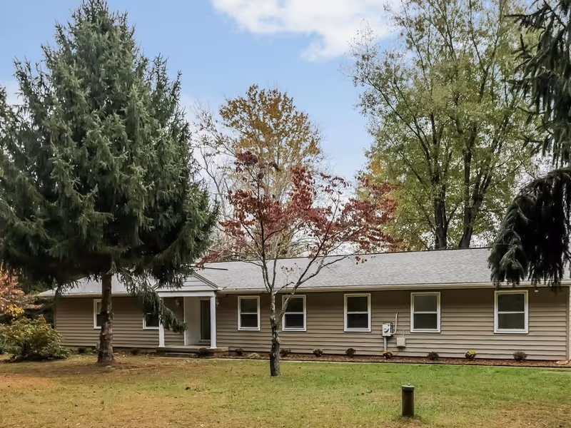 Single-story building with beige siding surrounded by trees with autumn foliage and a grassy lawn in front under a partly cloudy sky.