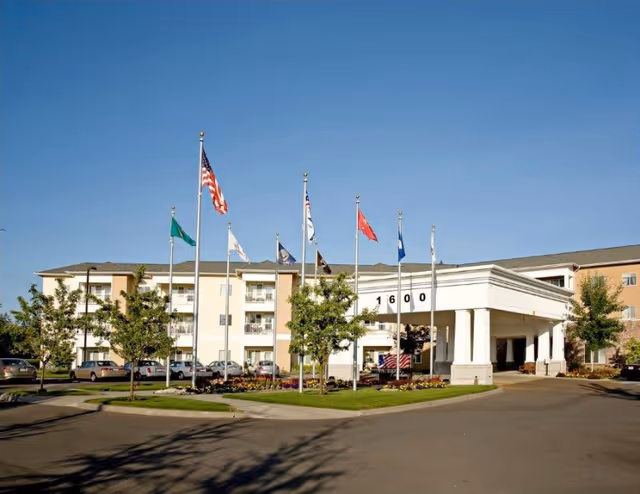 Exterior view of Patriot's Landing senior living facility showing a three-story building with a covered entrance. Several flagpoles with various flags are positioned in front of the building, along with landscaped greenery and parked cars.