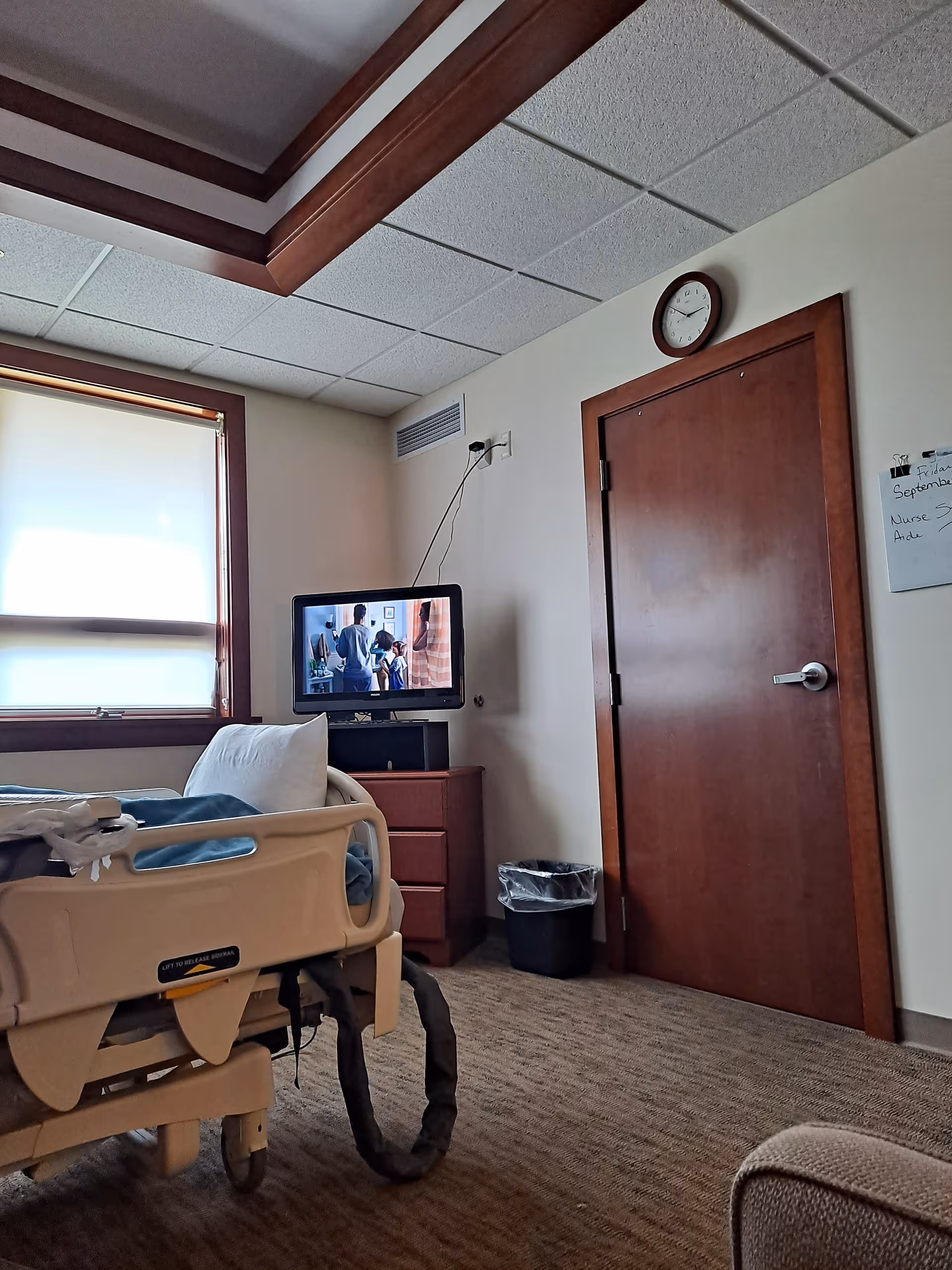 A hospital-style bed in a cozy resident room with a small TV and window.