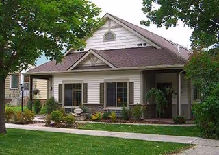 Single-story residential building with beige siding and a brown roof, surrounded by green trees and shrubs, with a concrete walkway leading to the entrance.