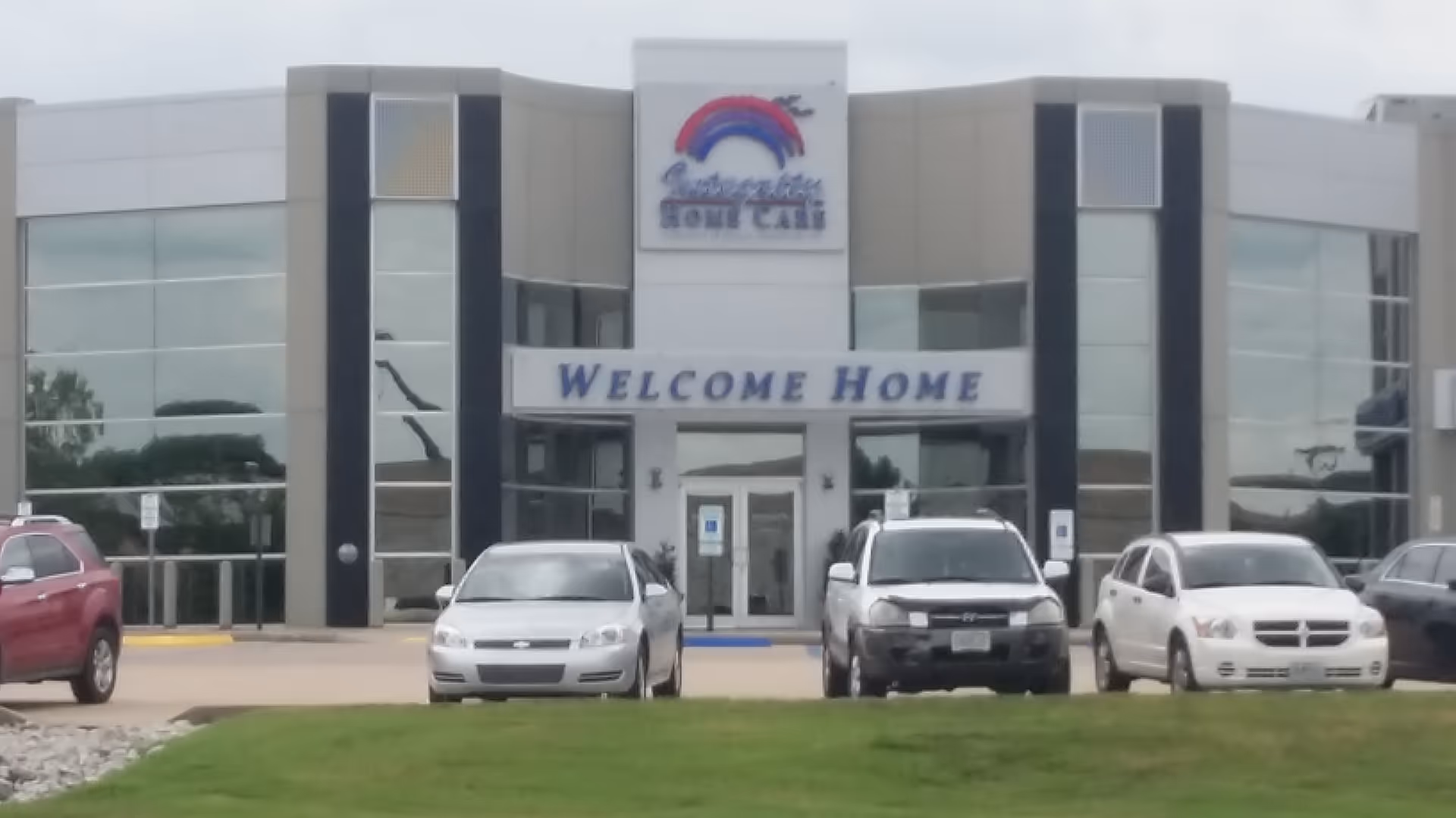 Modern facility with glass facade, entrance sign reading Welcome Home, and several parked cars.