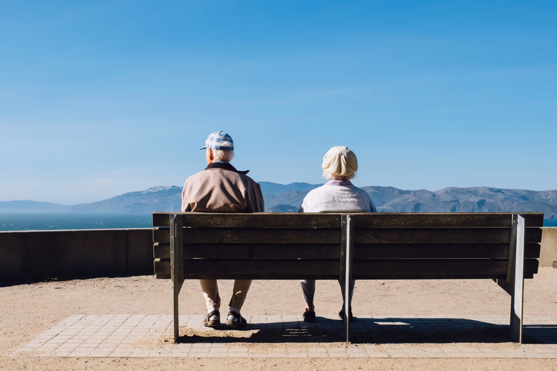 Two elderly people sit on a bench overlooking the ocean on an outdoor promenade.
