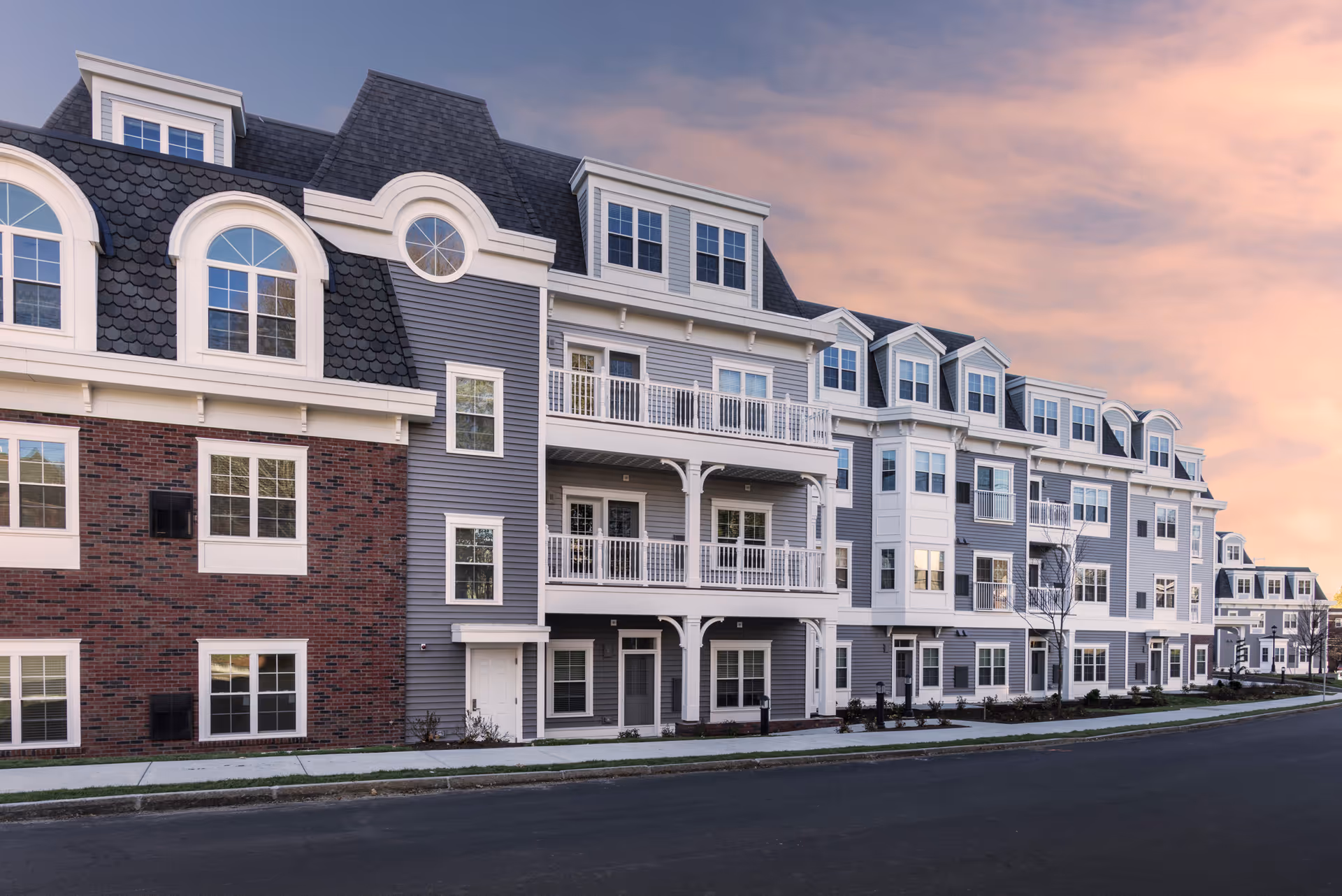 Street-facing exterior of a multi-story senior living building with balconies, dormer windows, and a brick-and-siding facade under a pink evening sky.
