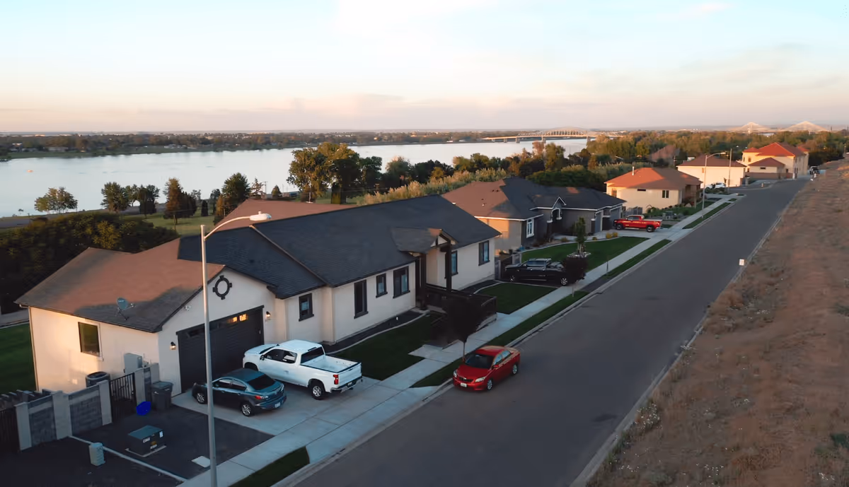 Aerial view of a residential street in Elysian Estates with several single-story houses, parked cars, a river in the background, and a bridge visible in the distance under a clear sky.