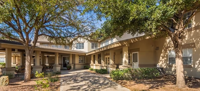 Courtyard entrance of a two-story senior living building with covered walkways, seating areas, and trees.