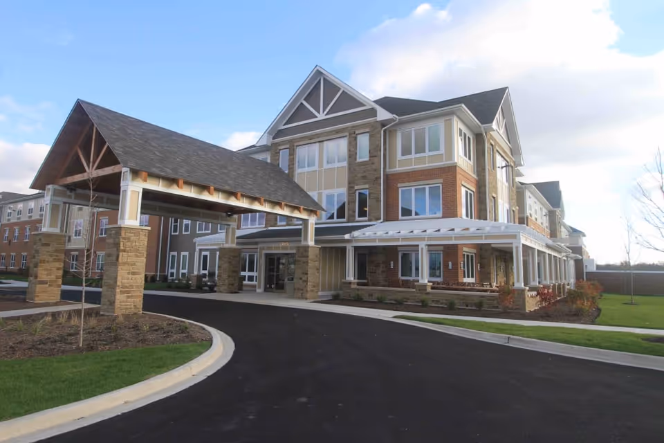 Exterior view of a multi-story senior living facility with a covered entrance supported by stone pillars, surrounded by landscaped grass and shrubs under a partly cloudy sky.