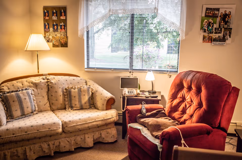 Cozy senior living room with a beige patterned sofa, red recliner, and a window behind.