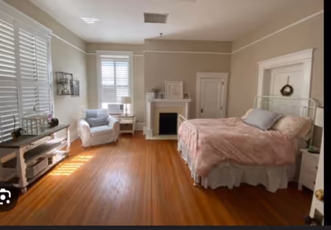 A spacious bedroom with a pink quilted bed, fireplace, and seating area.