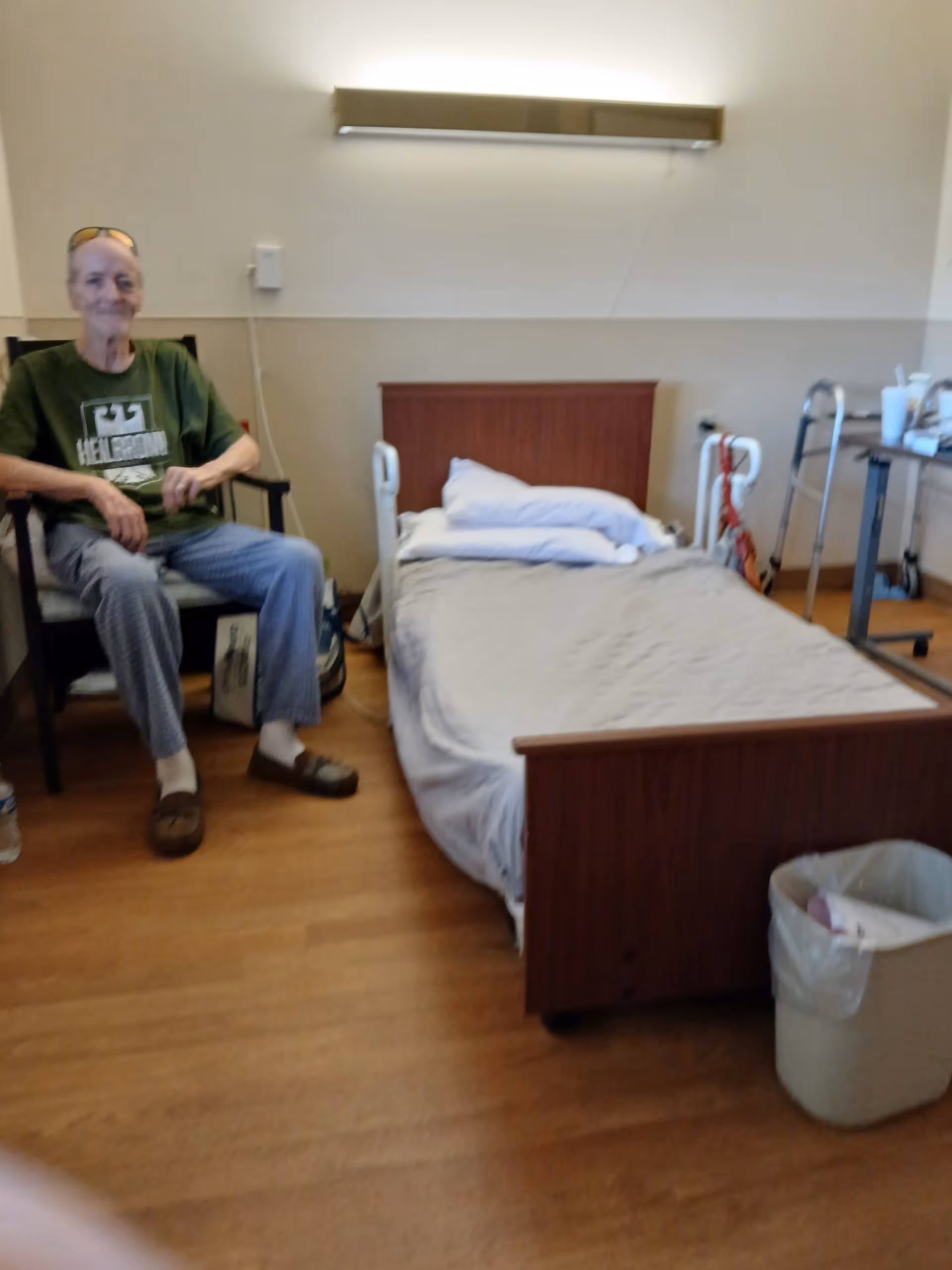 An elderly man sits in a chair beside a hospital-style bed in a senior living room.