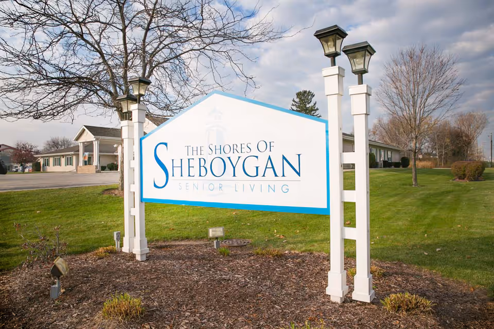 Exterior view showing The Shores of Sheboygan Senior Living sign, lawn, and buildings.