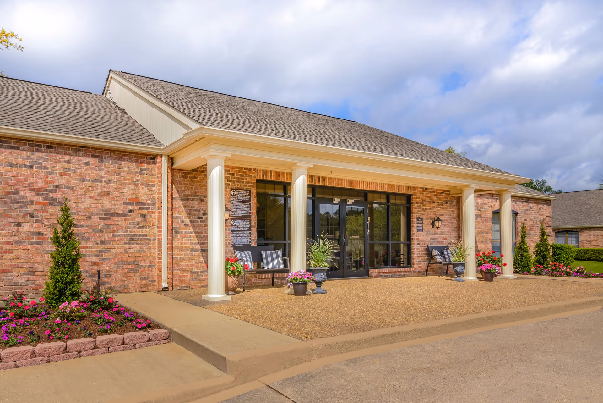 Front entrance of a brick nursing facility with white columns, glass double doors, potted flowers and benches.