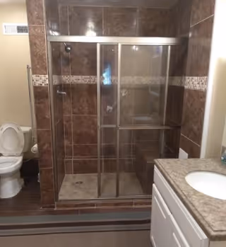 Bathroom with a glass-enclosed tiled shower, toilet to the left, and a vanity with sink to the right.