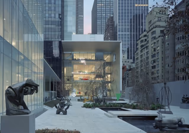 Modern outdoor courtyard surrounded by tall buildings, featuring abstract sculptures and a water basin.