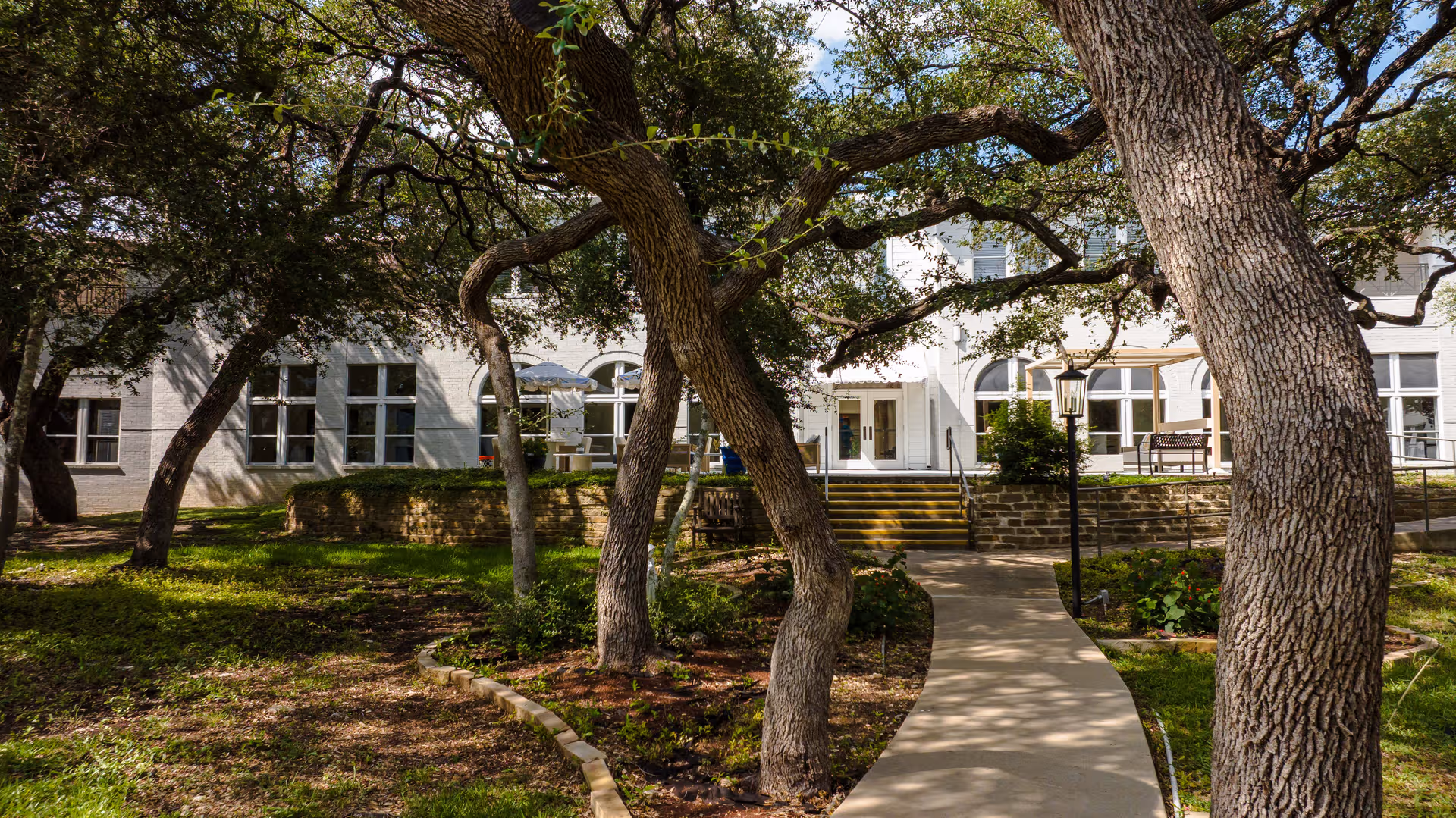 Curved walkway leads to a white senior living facility entrance framed by large trees.