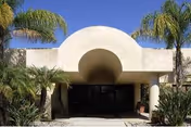 White stucco building front with arched entry, columns, and palm trees.